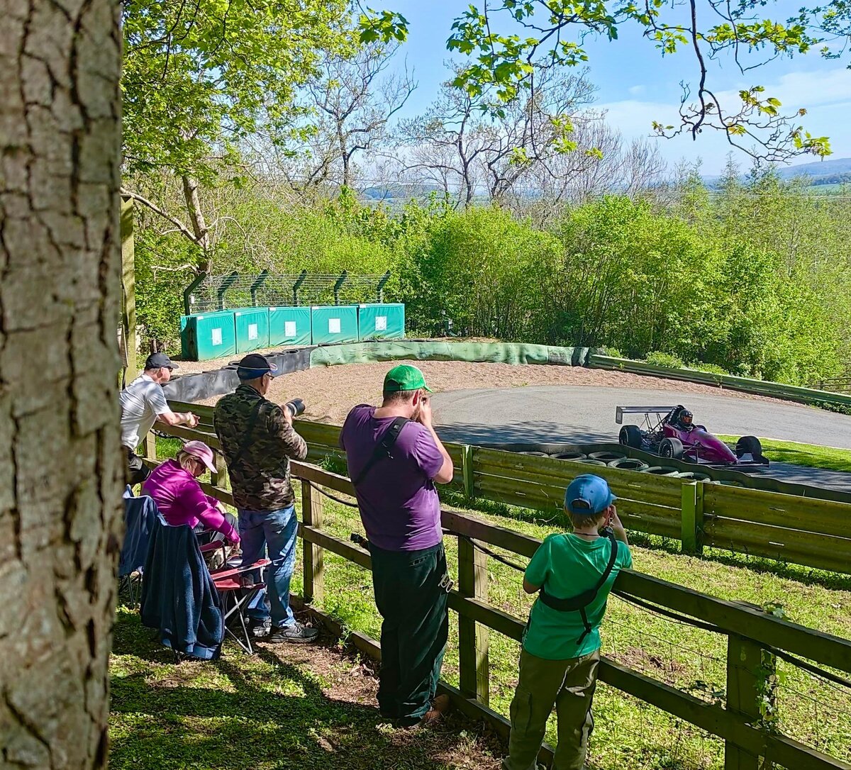 Photographers gathered at the fence of a motorsport circuit, karts visible on the track beyond.